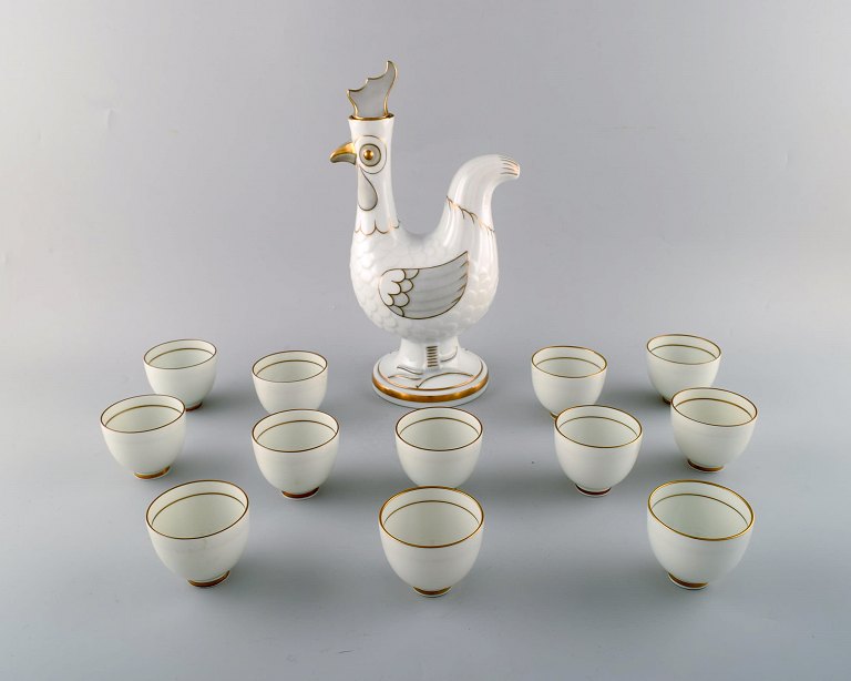 Bing & Grøndahl. Kande af porcelæn i form af hane, prop med hanekam. 12 
tilhørende krus af porcelæn med guldmalede detaljer.